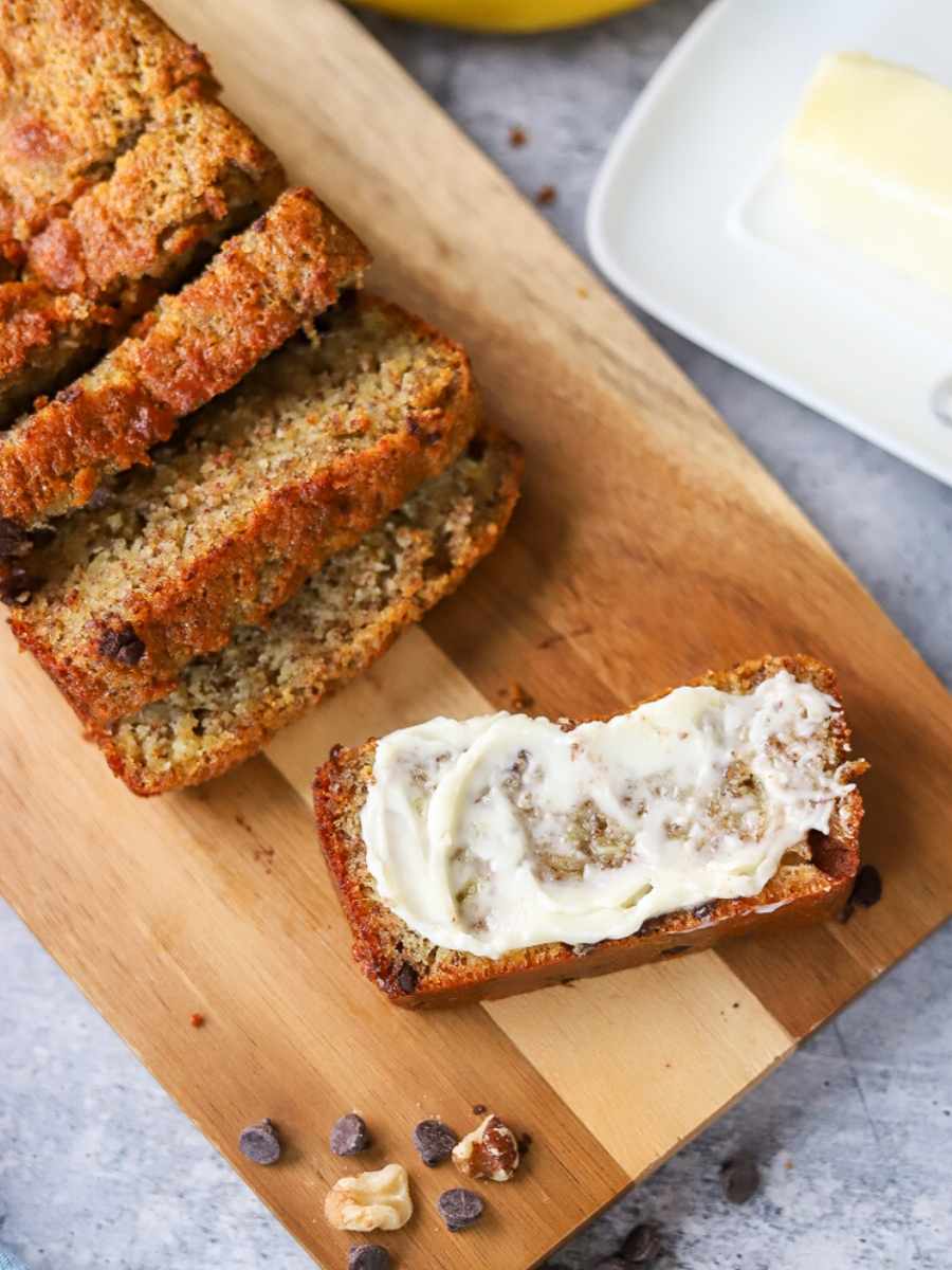 A loaf of banana bread on a cutting board with one sliced smeared with butter.