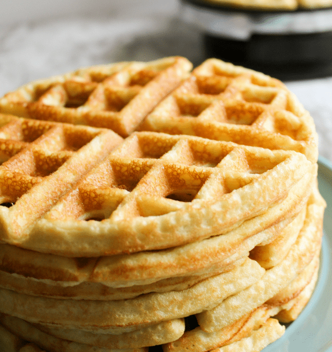 A tall stack of waffles with a waffle maker out of focus in the background