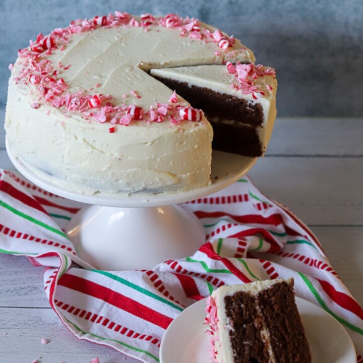 a round 2 layer cake on a white cake stand. The cake has white frosting and is topped with crushed peppermints. The cake has one slice missing.