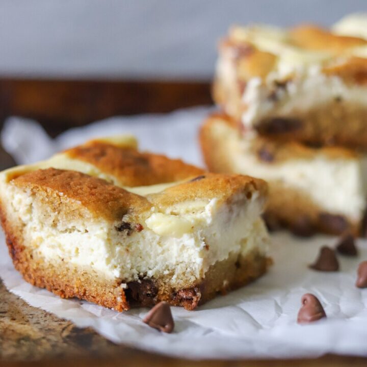 three chocolate chip cookie cheesecake bars on a piece of parchment paper with a bite taken from one