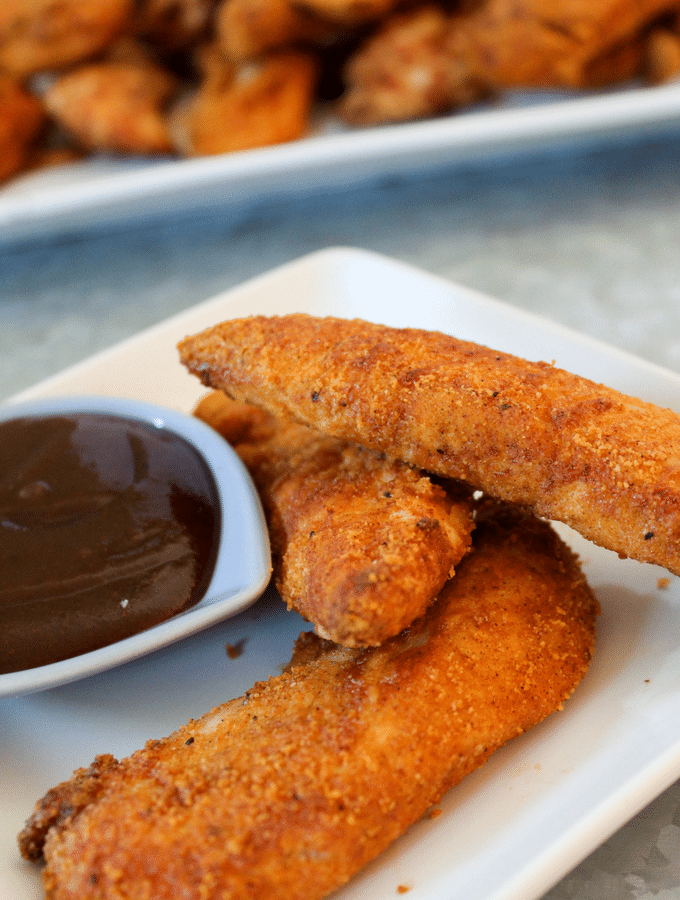 chicken tenders made with an almond flour breading stacked up on a white plate with sugar free barbecue sauce on the side.