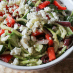 a round, white bowl filled with a salad of zucchini noodles, red peppers, feta cheese, olives, red onion and dressing.