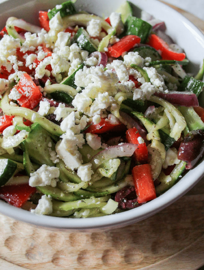 a round, white bowl filled with a salad of zucchini noodles, red peppers, feta cheese, olives, red onion and dressing.