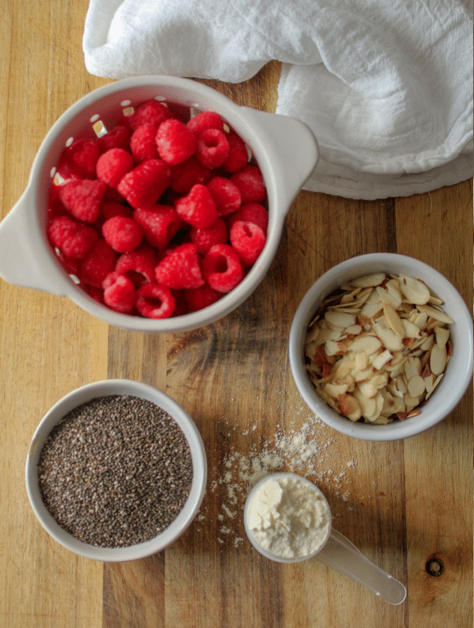 Ingredients for raspberry chia pudding on a  flat surface. This includes fresh raspberries, chia seeds, almonds, and vanilla protein powder.