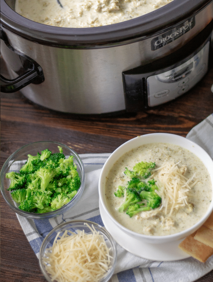 slow cooker chicken broccoli alfredo soup in a white bowl, with steamed broccoli and shredded Parmesan cheese on the side