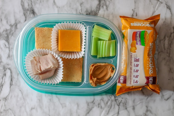 A 3 compartment food storage container with almond crackers, cheddar cheese, turkey and celery with peanut butter, plus a tangerine smart cake. 