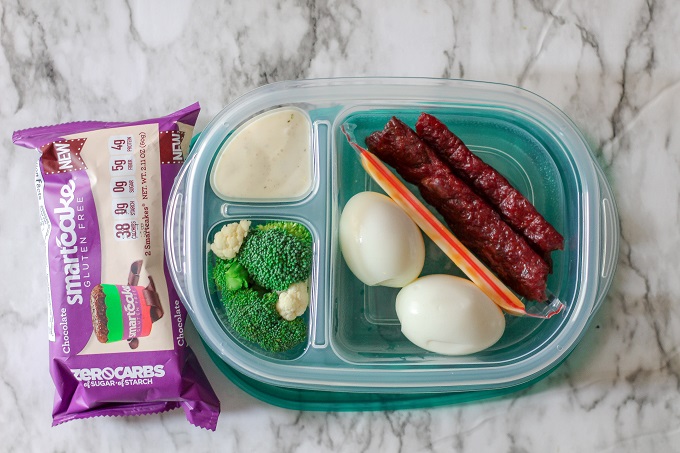 A 3 compartment food storage container with jerky, cheddar cheese, hard boiled eggs, broccoli and ranch, plus a chocolate smart cake. 