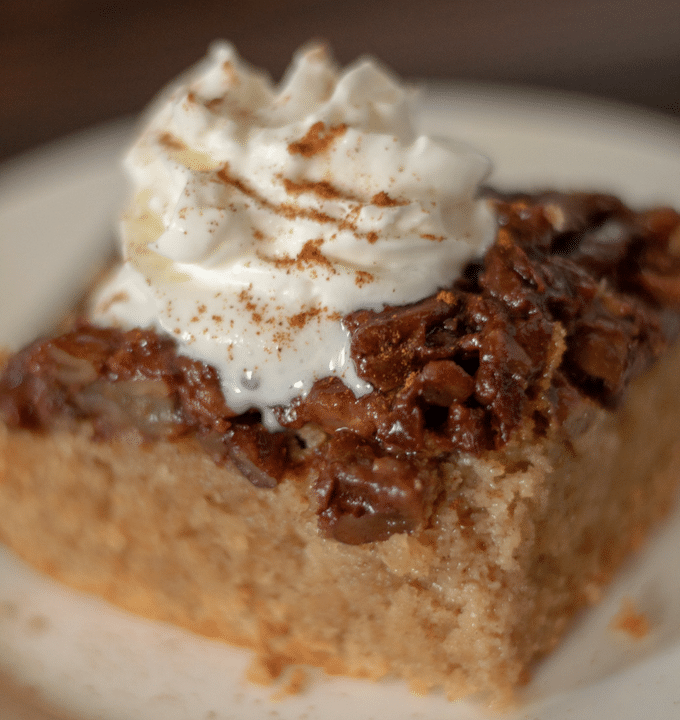 a square slice of upside down maple pecan cake topped with whipped cream, sitting on a round white plate.