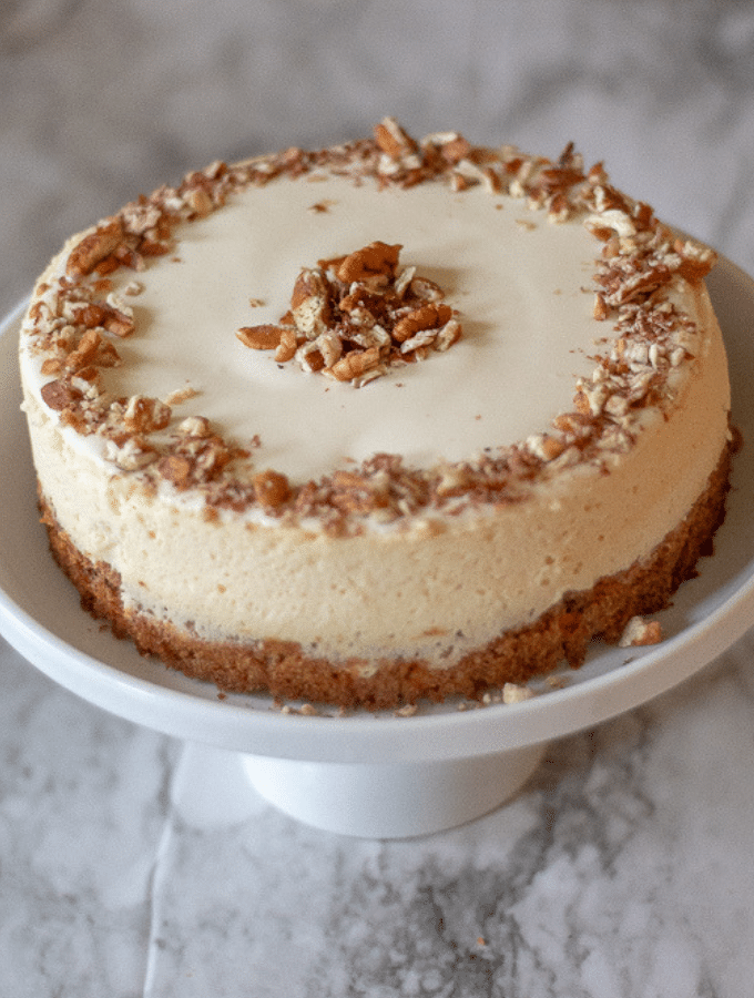 A keto carrot cake cheesecake on a white cakes stand garnished with chopped pecans
