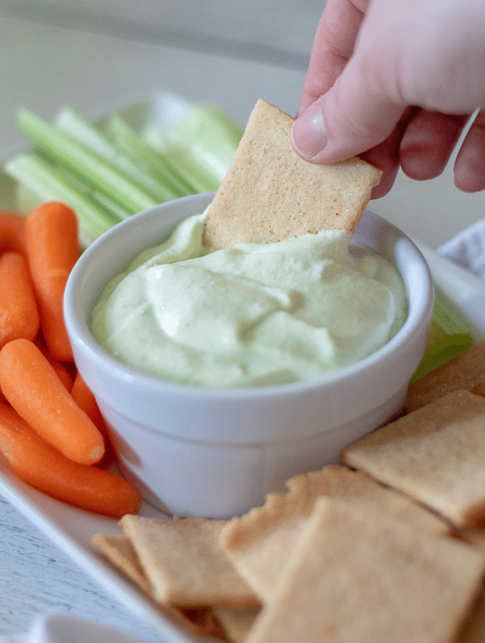 A White platter with a bowl of benedictine in the center surrounded by keto crackers, carrots and celery sticks. One butter cracker is being dipped into the benedictine
