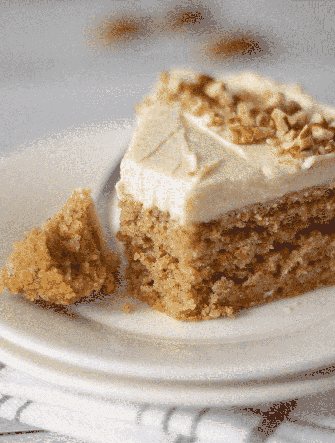 A slice of carrot cake made with almond flour and topped with cream cheese frosting on a white plate with one bite taken. 