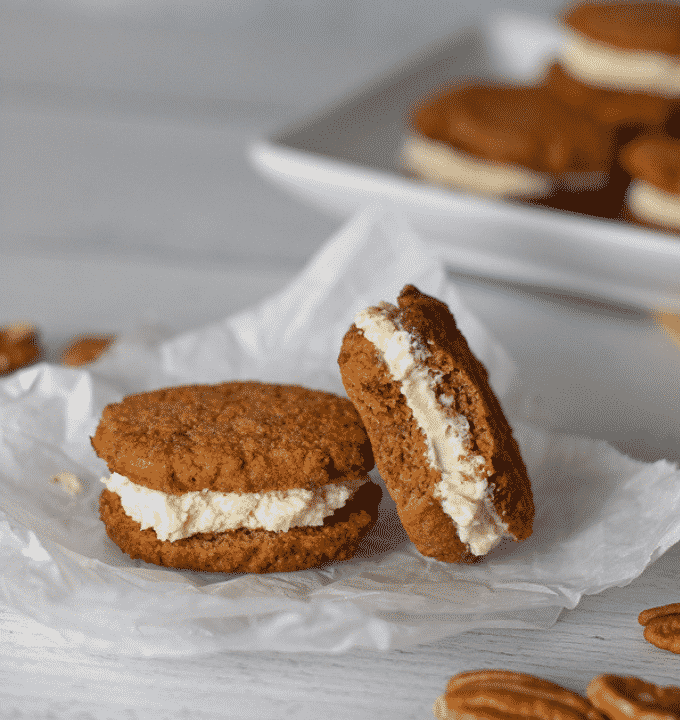 two Keto carrot cake whoopie pies on a piece of parchment with a scattering of pecans and a tray with more .whoopie pies in the backgound