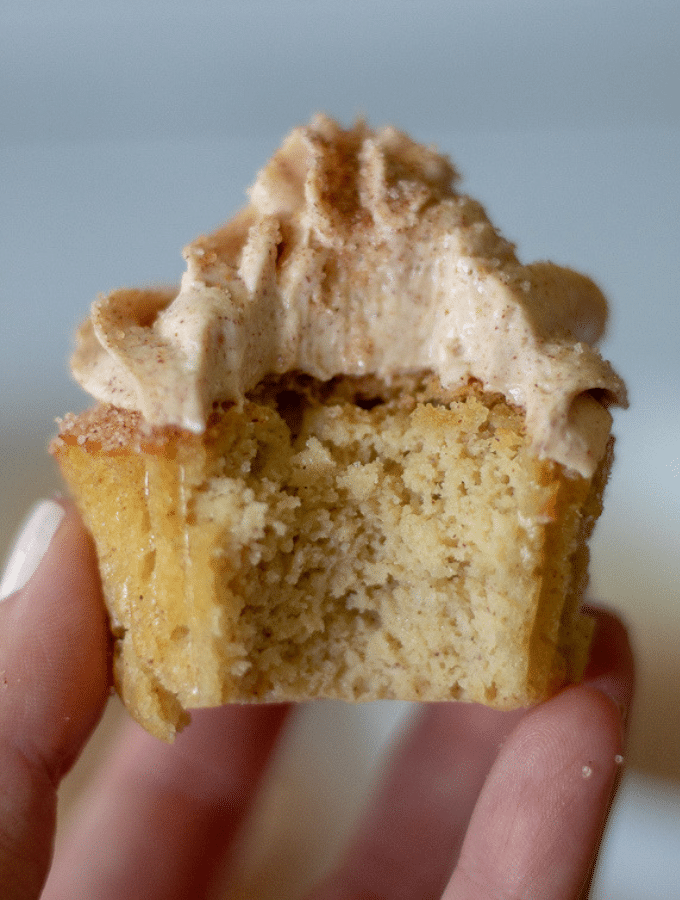 A single cinnamon sugar coated cupcake topped with cinnamon cream cheese icing. One bite has been taken and the cupcake is being help up.