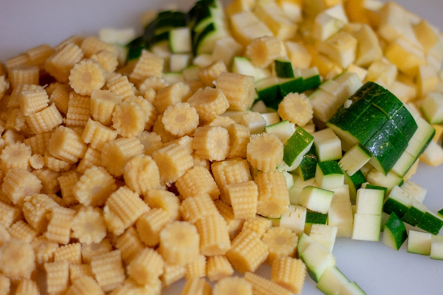 Diced baby corn, zucchini, and yellow squash on a white cutting board in preparation for keto Mexican street corn salad