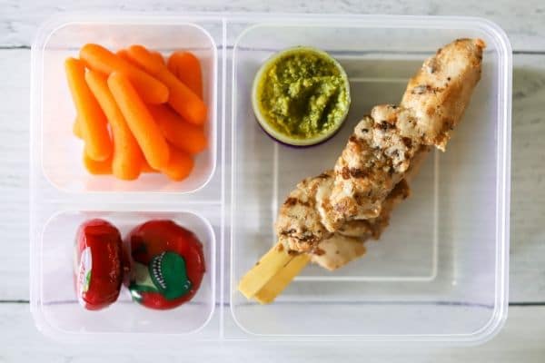 two grilled chicken skewers with pesto dipping sauce. carrots, and cheese in a lunch box.