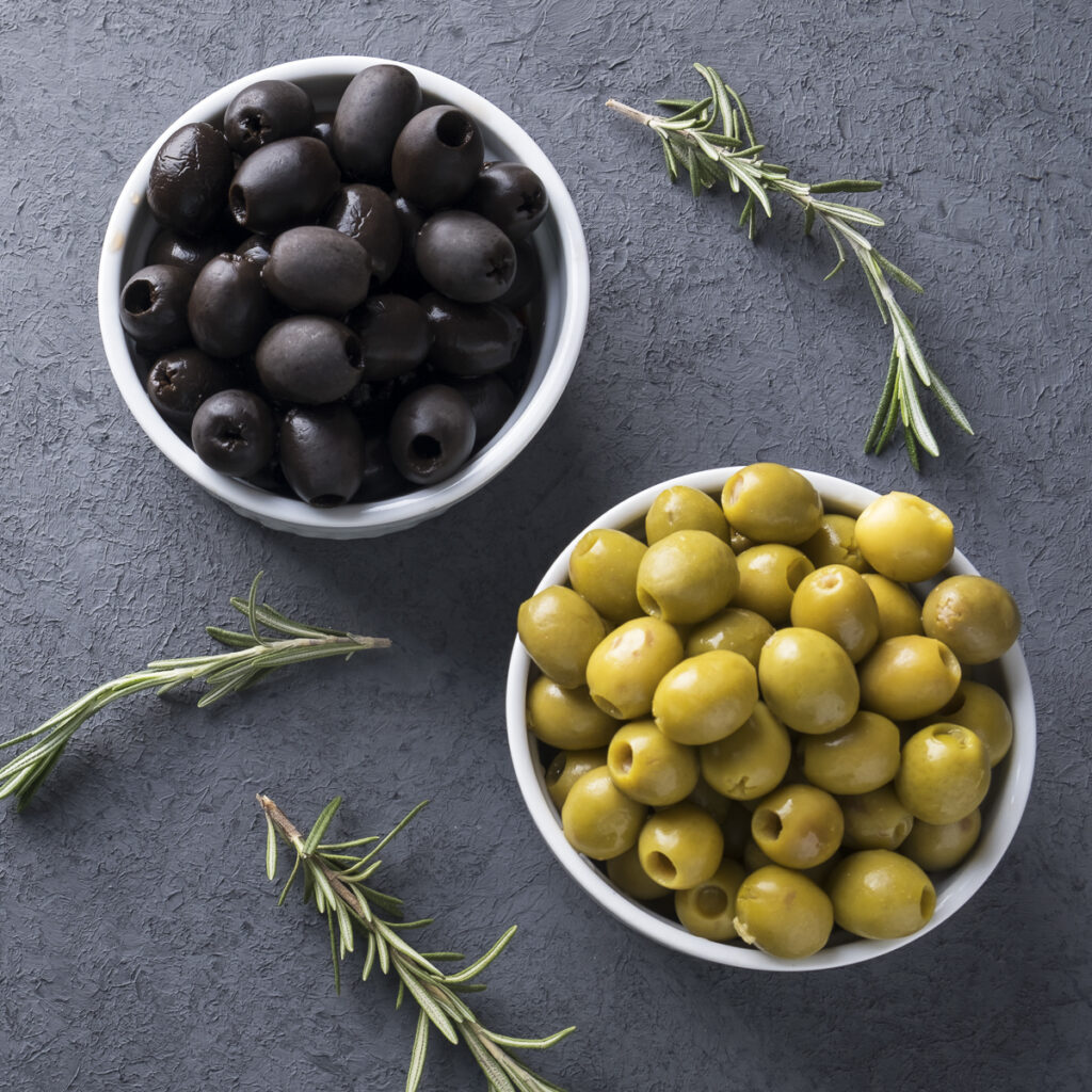 two white bowls of olives, one with green olives and the other with black olives. Rosemary sprigs are off to the side. 
