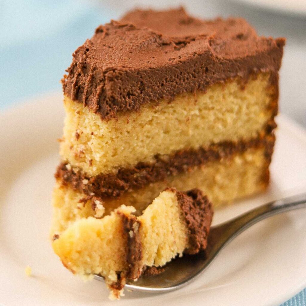 A single slice of keto yellow cake with chocolate frosting on a white plate with one bite taken