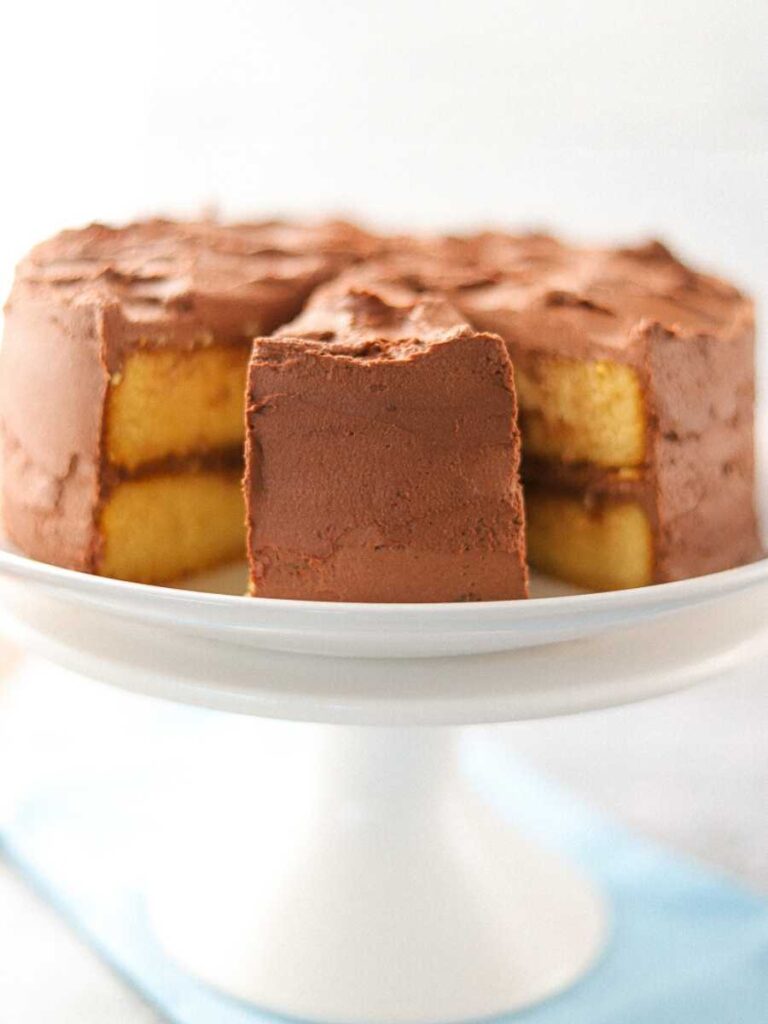 A yellow cake with chocolate frosting on a white cake stand with two slices removed