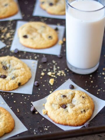 chocolate chip cookies on individual parchment squares, with scatted crumbs and chocolate chips plus a glass of milk off to the side.
