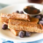a plate full of homemade french toast sticks with blueberries and a silver cup of sugar free maple syrup.