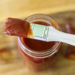 A small mason jar full of homemade barbecue sauce with a pastry brush covered in sauce balanced on top