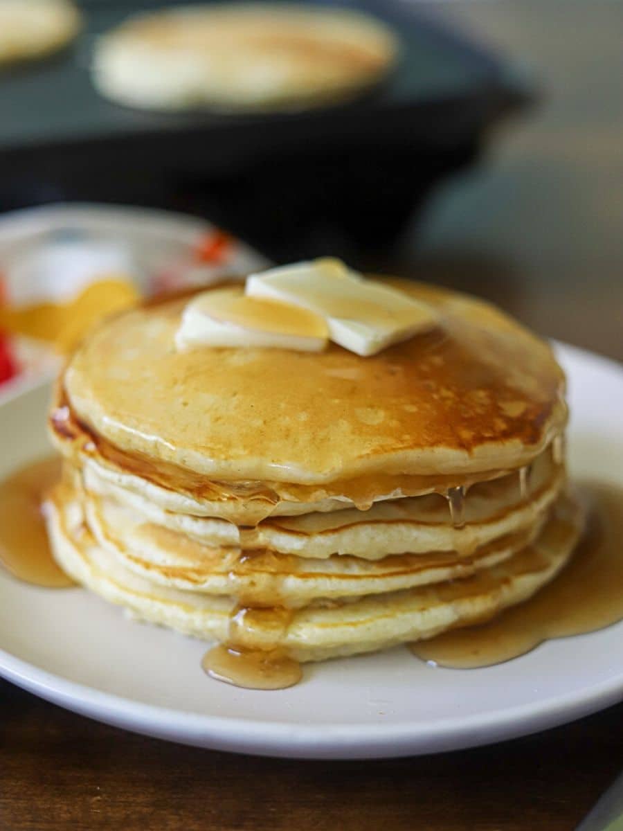 A stack of pancakes on a white plate, topped with two pats of butter and maple syrup.