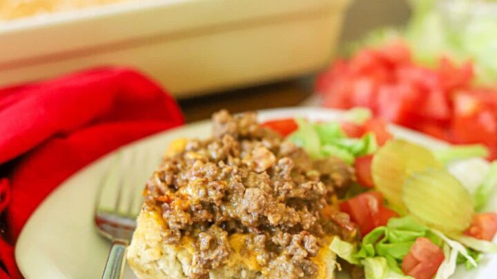 a serving of cheseburger casserole on a white plate with chopped tomatoes, lettuce, and pickles on the side