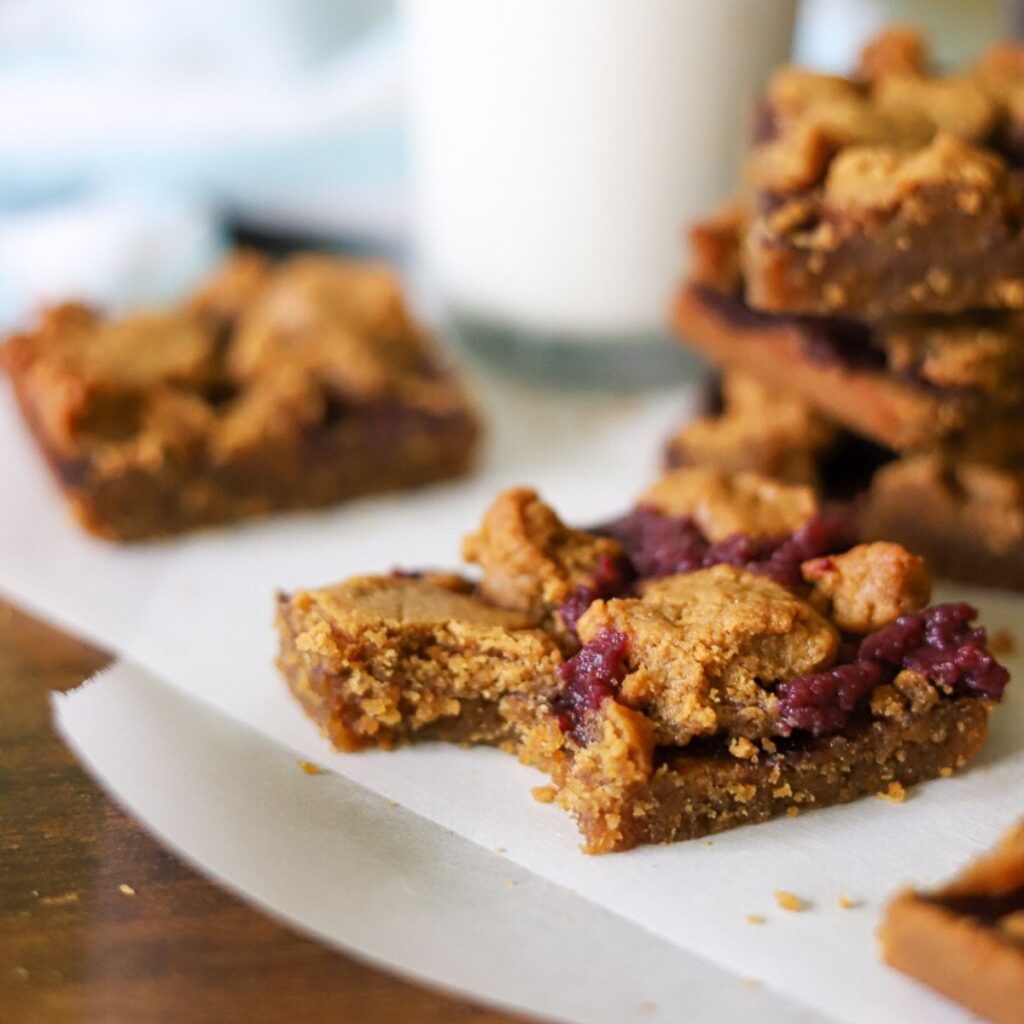 peanut butter and jelly bars on a piece of parchment paper. One has a bite taken and a glass of milk is in the background.