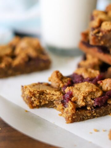 peanut butter and jelly bars on a piece of parchment paper. One has a bite taken and a glass of milk is in the background.