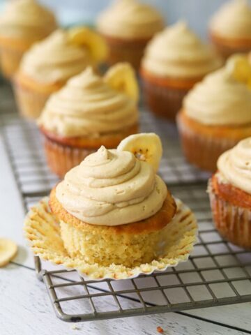 gluten free banana cupcakes on a wire cooling rack. The cupcakes are topped with peanut butter frosting and a single banana chip on each cupcake.