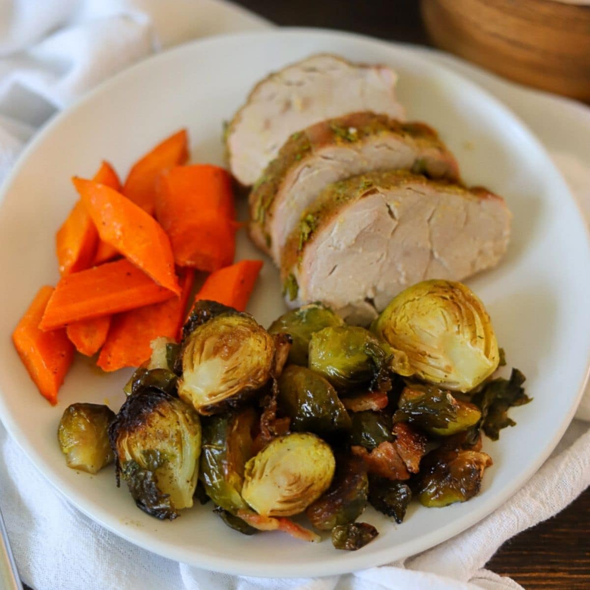 a white plate filled with sliced pork tenderloin, roasted carrots, and brussels sprouts