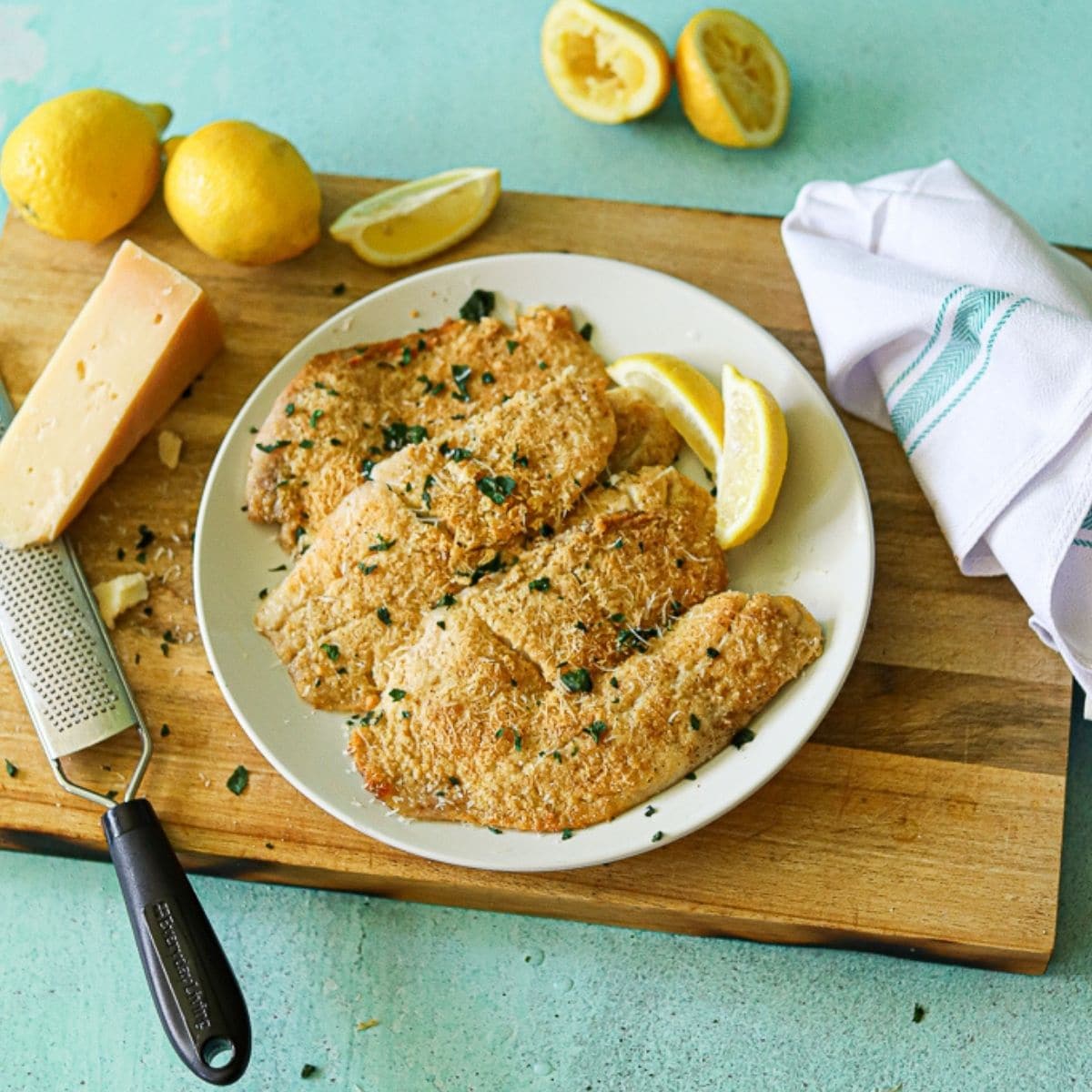 Baked tilapia on a white plate with lemon and fresh parmesan cheese off to the side.