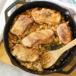 a cast iron skillet full of cauliflower rice topped with pork chops. A wooden spoon os resting on the side.
