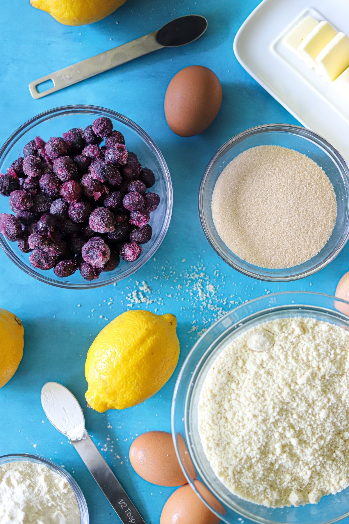 Lemons, blueberries, Almond flour, eggs, sweetener, butter, and vanilla on a flat surface.