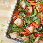 Chicken, snow peas, red pepper, carrots, broccoli, and sesame seeds cooked together on a sheet pan.