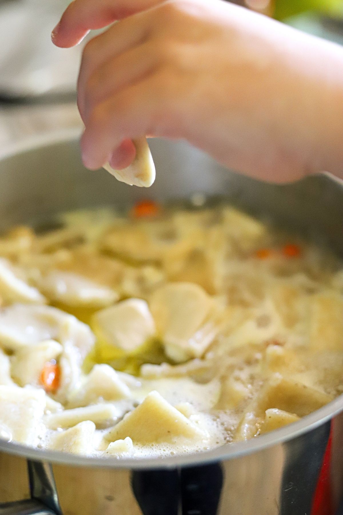 A child's hand dropping a rolled dumpling into a pot of boiling chicken and dumplings