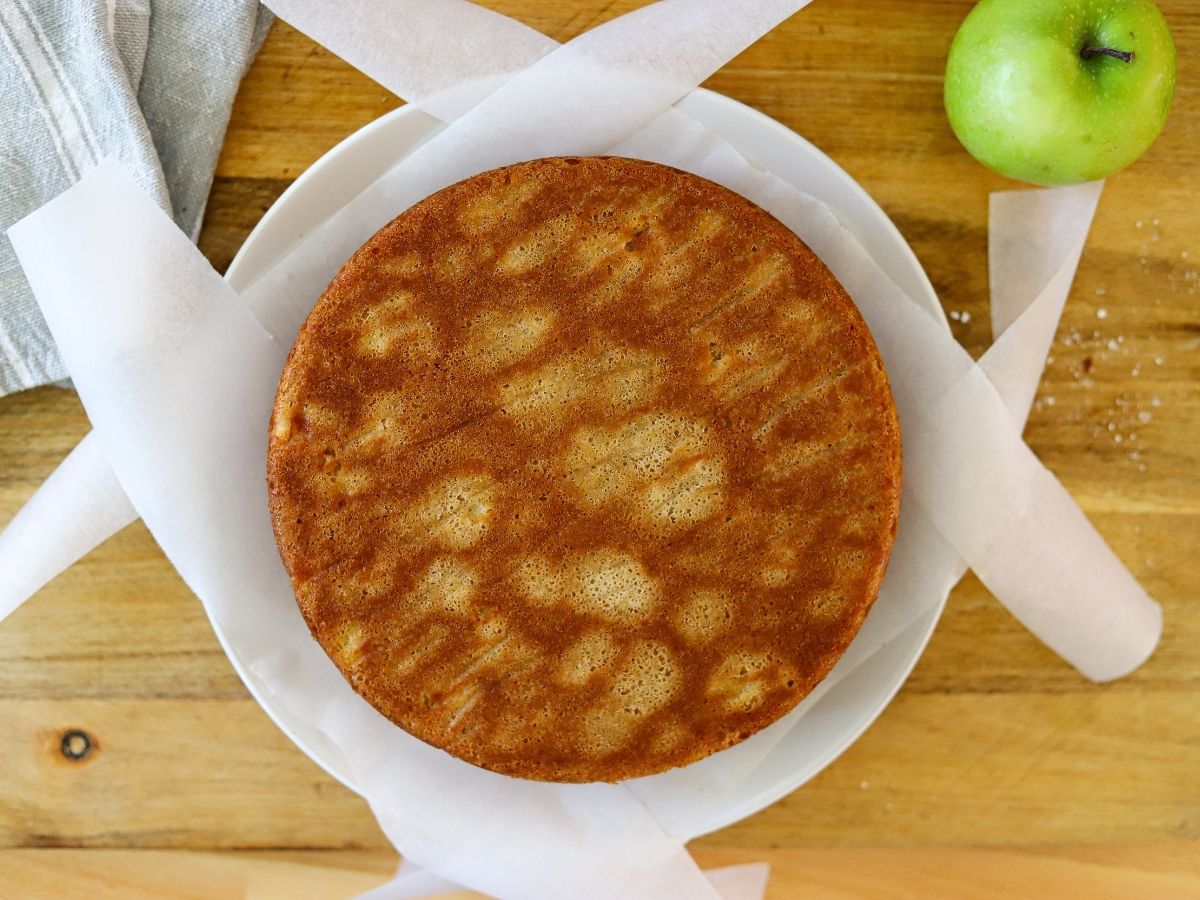 A cooked apple cake on a serving plate with strips of parchment paper under the edges.