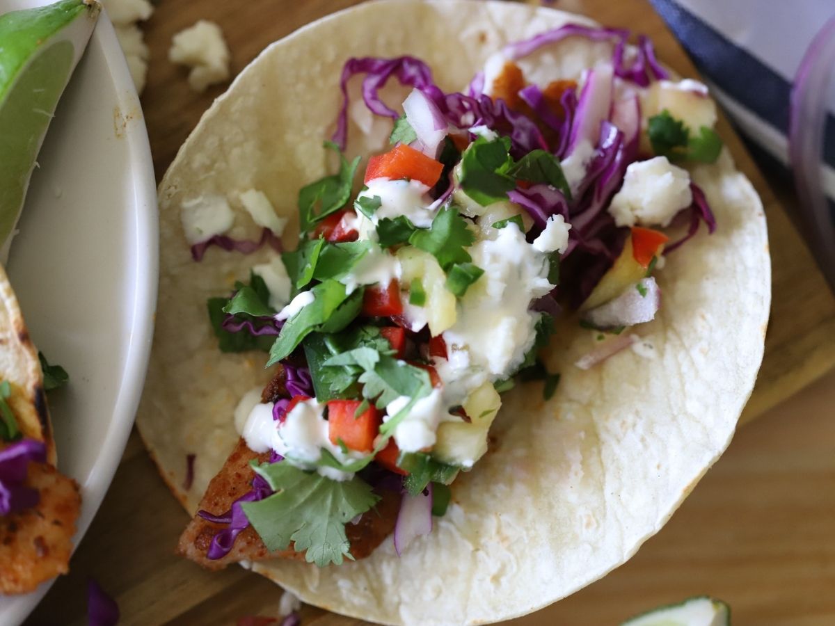 a fish taco topped with red cabbage, cilantro, queso fresco, pineapple, and a lime sauce