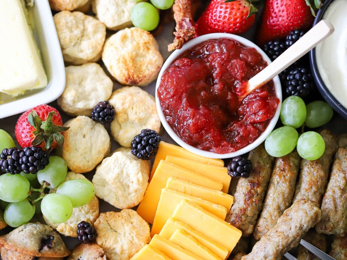 an overhead image of a breakfast charcuterie board with biscuits, sausage links, cheese, jam, and berries