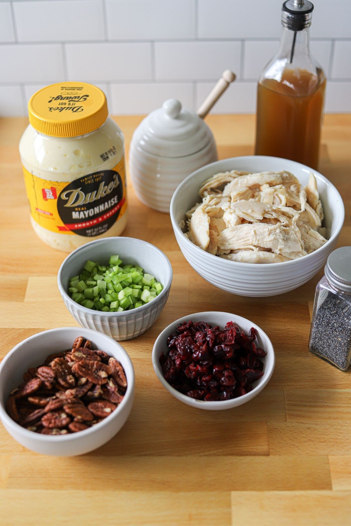 Mayonnaise, celery, pecans, pulled chicken, dried cranberries, vinegar, honey, and poppy seeds laid out on a wooden counter to make chicken salad