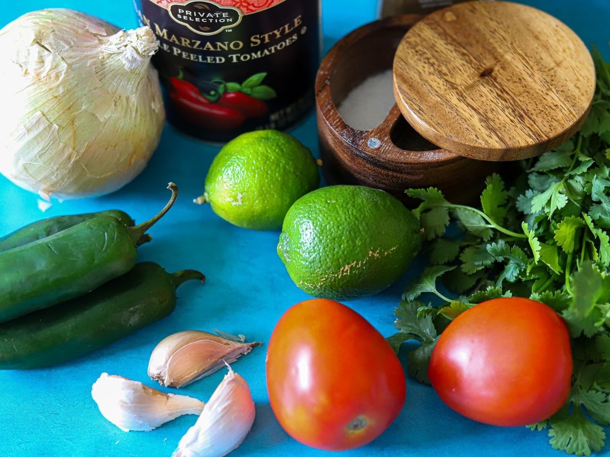 ingredients for an easy homemade salsa including canned whole tomatoes, onion, jalapeños, limes, cilantro, garlic, roma tomatoes, and salt and pepper