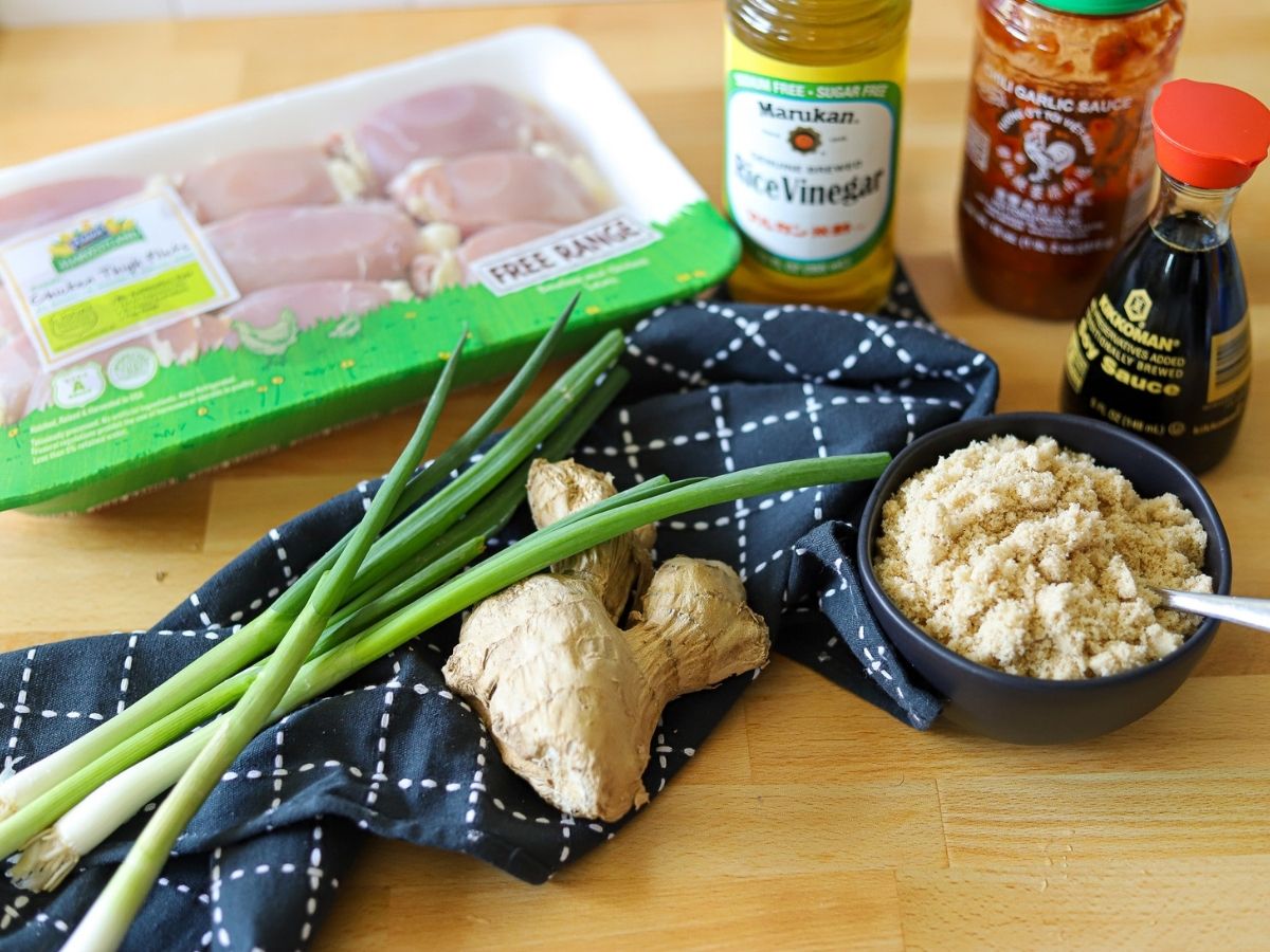 Ingredients for sticky Asian glazed chicken thighs on a wooden surface. The ingredients include boneless, skinless chicken thighs, ginger,  green onions, brown sugar, garlic chili sauce, rice wine vinegar, and soy sauce