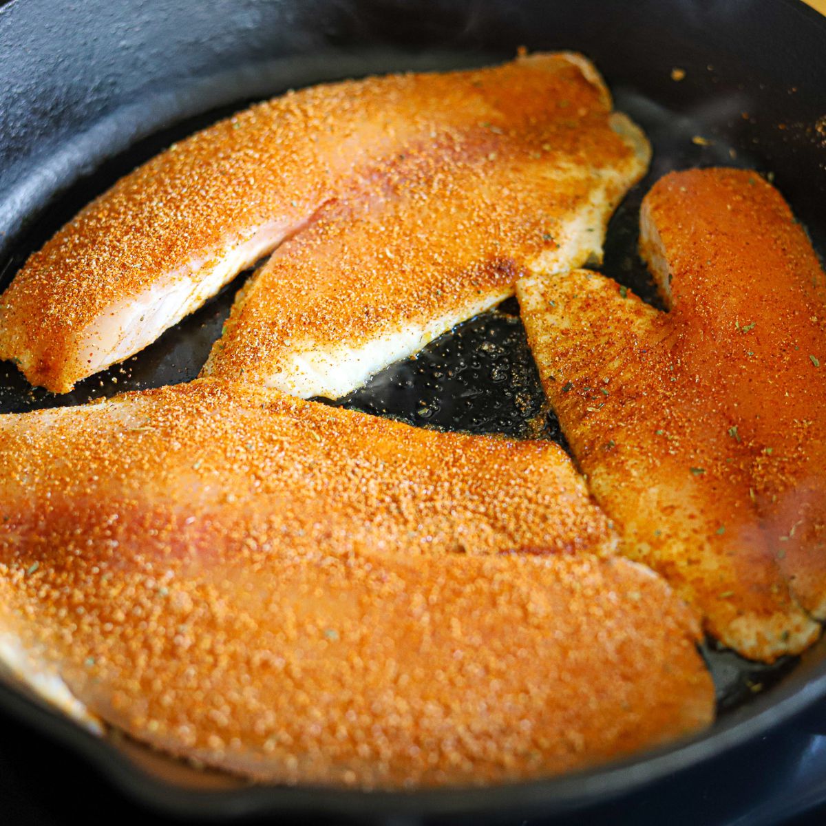 tilapia fillets seasoned with blackening seasoning, cooking in a cast iron skillet.