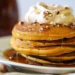 a stack of sweet potato pancakes topped with maple syrup, toasted pecans, and whipped cream