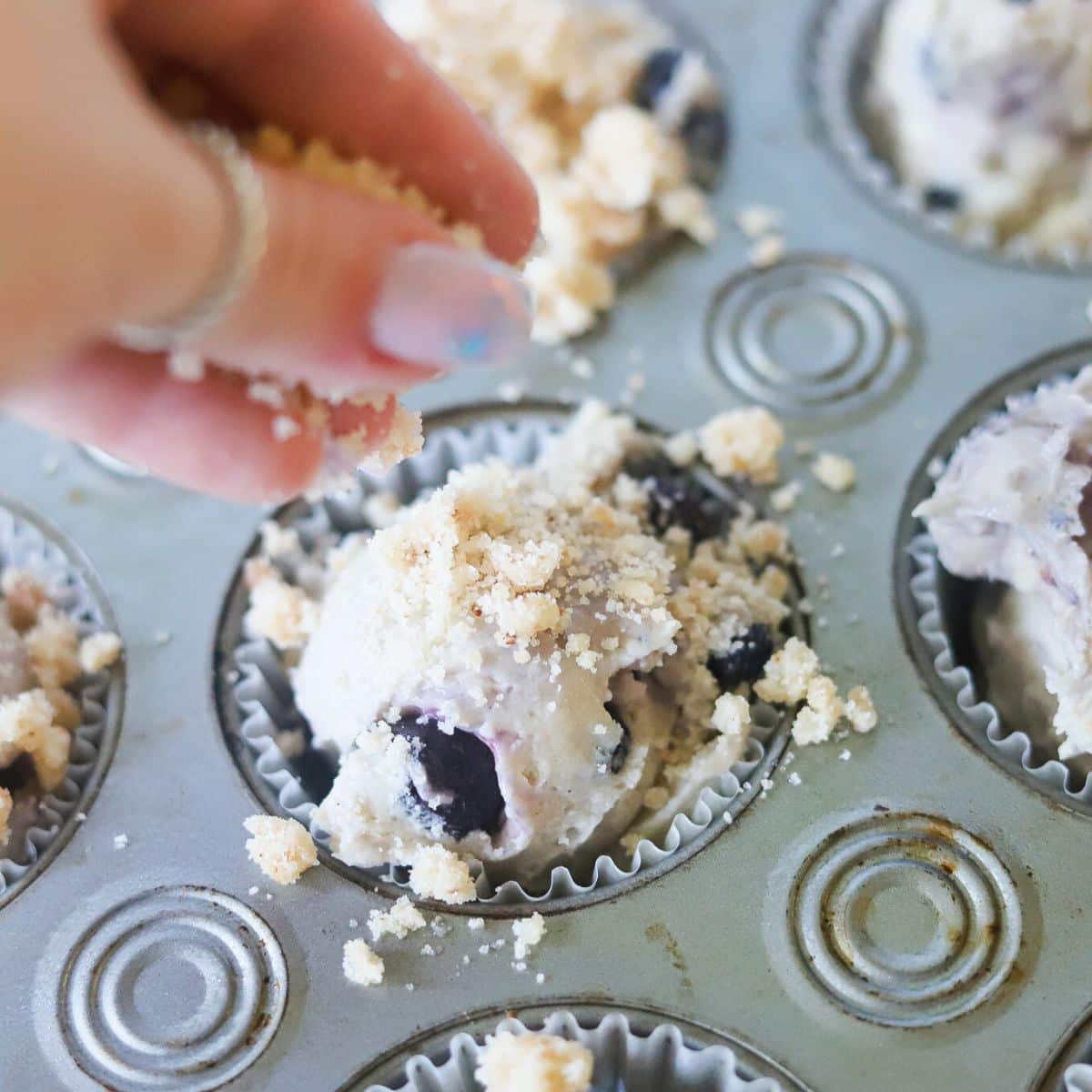 blueberry muffin batter in a muffing tin being topped with a crumble. 