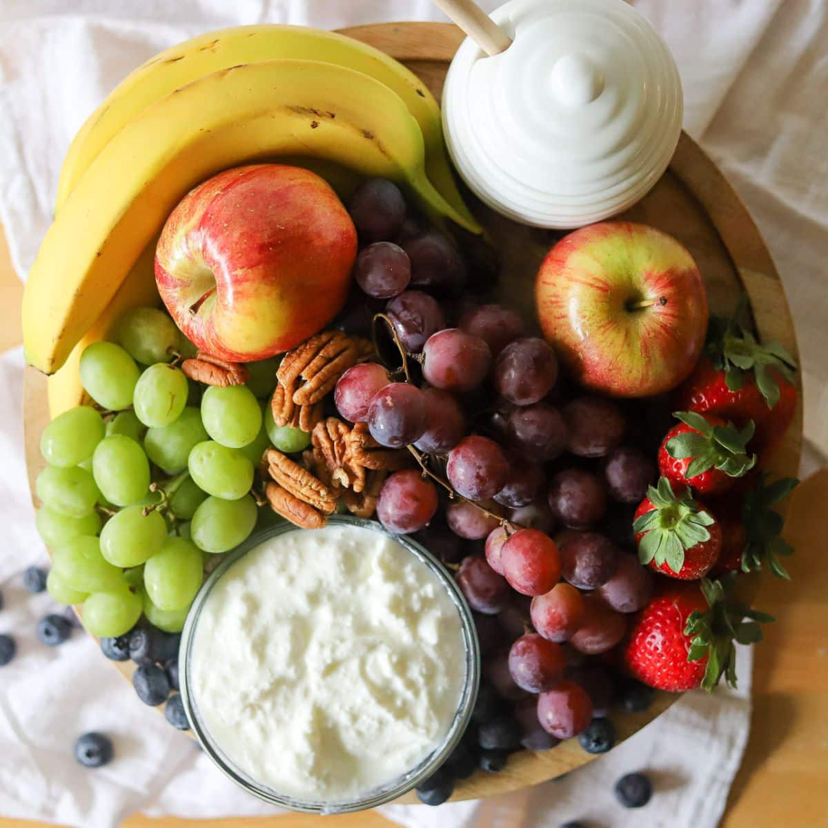 ingredients for a delicious breakfast fruit salad including bananas, apples, grapes, blueberries, strawberries, pecans, yogurt, and honey.