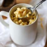 a coffee mug full of macaroni and cheese. One bite is being lifted from the mug in a black spoon.