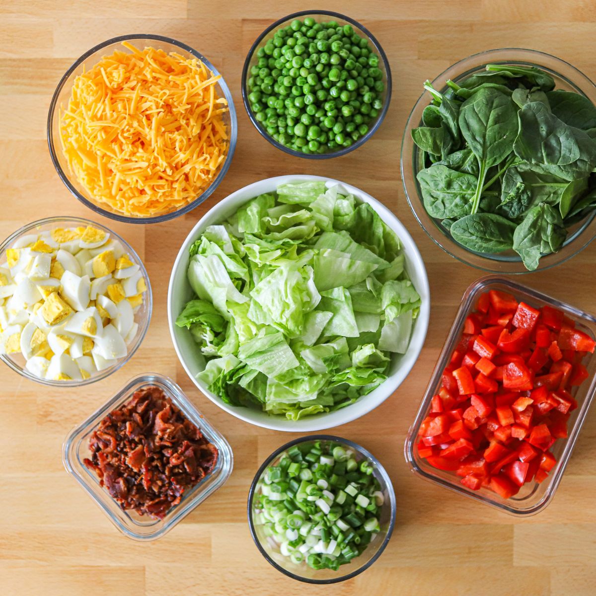 8 bowls of various sizes filled with the ingredients for a layered salad including baby spinach, iceberg lettuce, peas, cheddar cheese, hardboiled eggs, bacon, red bell pepper, and green onion.
