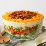 A glass bowl filled with a layered salad topped with shredded cheese and chopped bacon.