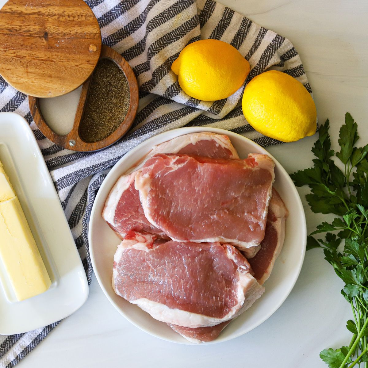 ingredients for lemon pepper pork chops including a plate full of pork chops, fresh lemons, salt, pepper, butter, and fresh parsley.